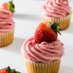 A group of cupcakes on white topped with strawberry cream cheese frosting topped with a fresh strawberry