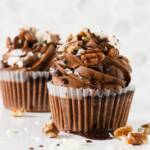 Two German chocolate cupcakes on a counter decorated with chocolate frosting swirls, coconut flakes, pecans, and drizzled with chocolate syrup