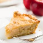 slice of apple custard pie on a white plate with a fork and apples in the background