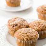 Three cinnamon muffins on a white plate with a muffin in the background