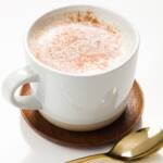 White mug of cinnamon milk on a wood coaster with a gold spoon in front of it