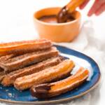 Blue plate with a stack of churros and a churros being dipped into chocolate sauce in the background